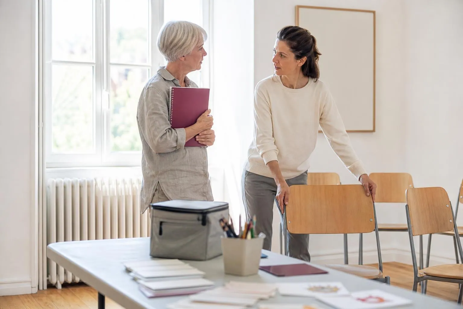 Deux consultantes debout près d’une grande fenêtre dans une salle associative claire, carnets à la main, rangeant des chaises disposées en cercle